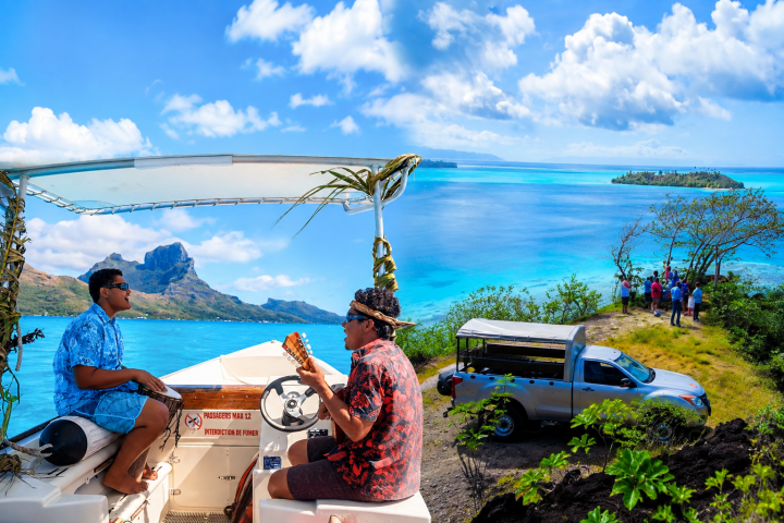 Two musicians on a boat near turquoise water, with people and vehicle on a nearby hill.